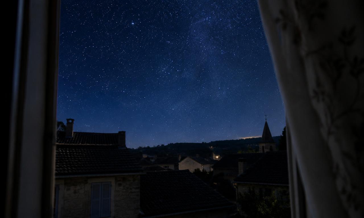 Le ciel étoilé au-dessus des toits d'un petit village, aperçu depuis une fenêtre entrouverte.